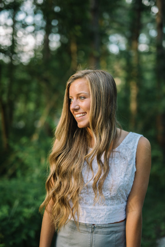 A young woman with long wavy blonde hair, wearing a white sleeveless top, smiles while standing outdoors in a sunlit, green forest. by Justin Salem Meyer
