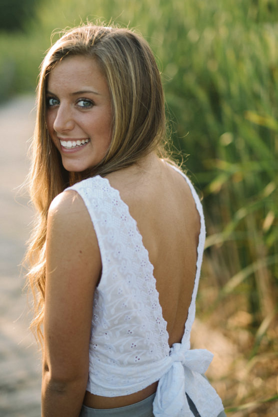 A young woman with long hair smiles over her shoulder, wearing a white, backless, lace top with a bow at the lower back. She stands outdoors near tall green grass, in soft, natural light. by Justin Salem Meyer