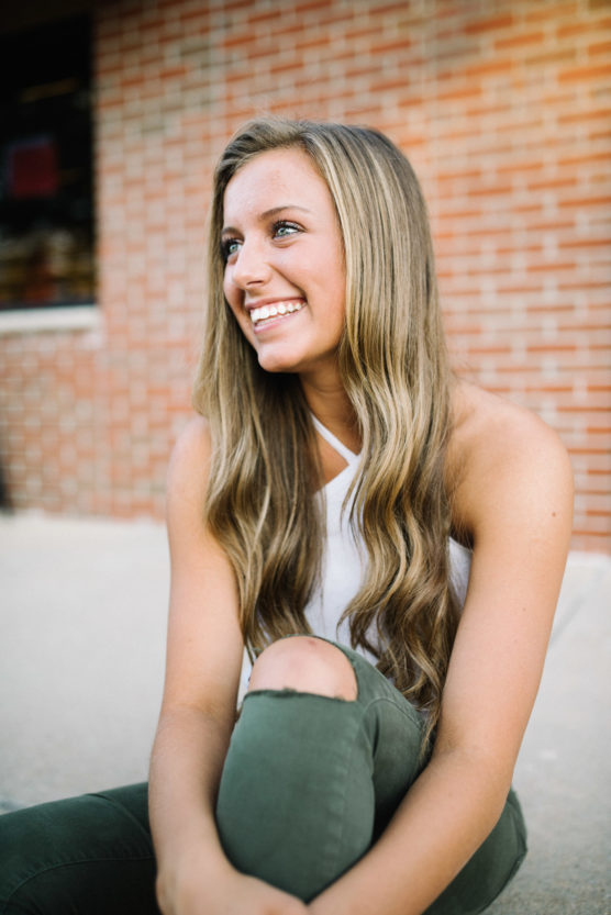 A young woman with long, wavy blonde hair, wearing a white sleeveless top and olive green pants with a ripped knee, sits outdoors and smiles, with a brick wall in the background. by Justin Salem Meyer
