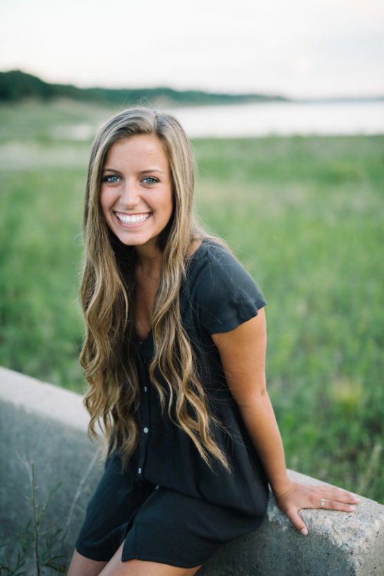 A young woman with long wavy hair, wearing a short-sleeved black dress, sits on a concrete ledge outdoors and smiles at the camera with a blurred grassy field and water in the background. by Justin Salem Meyer