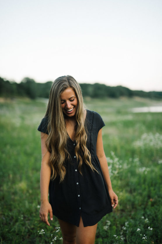 A young woman with long wavy hair wearing a short-sleeved black dress smiles and looks down while standing in a green field with wildflowers. Trees and a river are visible in the background. by Justin Salem Meyer
