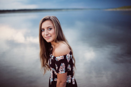 A young woman with long hair wearing a floral off-the-shoulder dress stands by a calm lake, smiling softly at the camera with a serene sky and water in the background. by Justin Salem Meyer