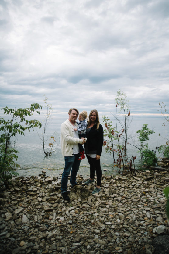 A family of three stands on a rocky shoreline by a lake, with overcast skies and sparse trees in the background. The adult on the left holds a child, while the other adult stands beside them, all smiling at the camera. by Justin Salem Meyer