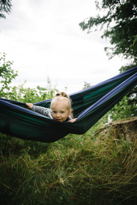 A young child with blonde hair and a striped shirt smiles while lying in a blue and green hammock outdoors, surrounded by grass, bushes, and trees. by Justin Salem Meyer