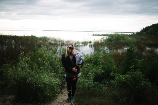 A woman wearing sunglasses stands on a narrow path surrounded by green bushes, holding a child in striped clothes. Behind them is a calm body of water and a cloudy sky. by Justin Salem Meyer