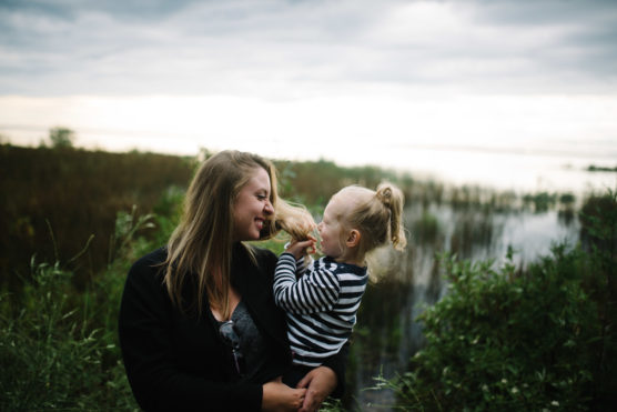 A woman smiles as a young child in her arms playfully pulls her hair. They are outdoors near water and tall grass, with a cloudy sky in the background. by Justin Salem Meyer