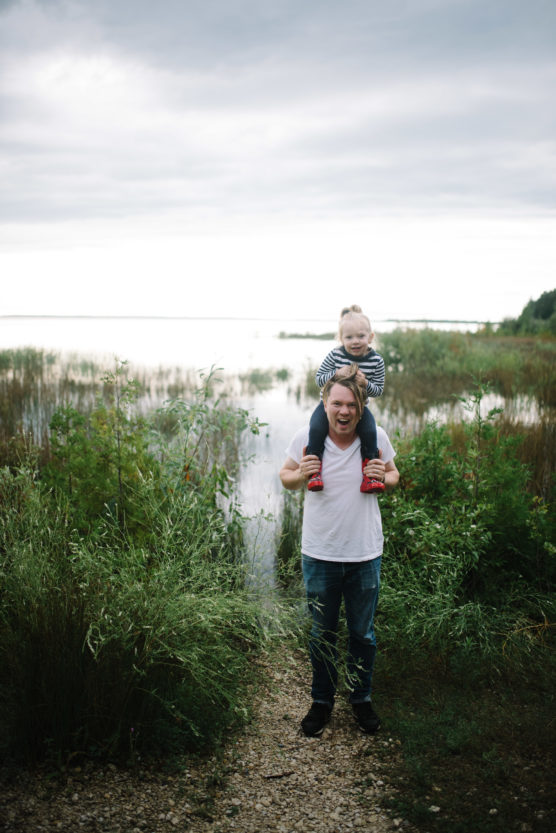A man stands on a gravel path by a lake with green plants on either side, smiling as he carries a young child on his shoulders. The child wears a striped outfit and red shoes. The sky is overcast and the water is calm. by Justin Salem Meyer