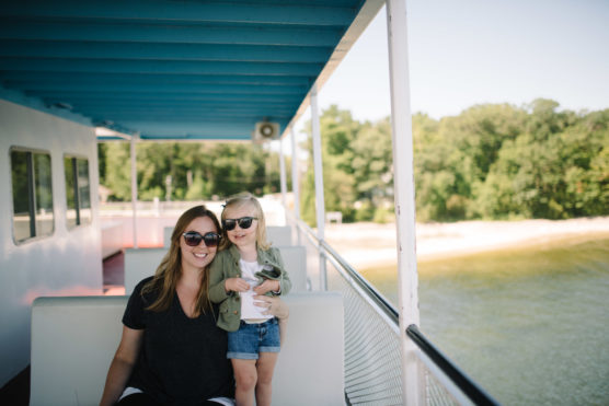 A woman and a young girl, both wearing sunglasses, sit together and smile on a boat with a blue roof. Trees and water are visible in the background on a sunny day. by Justin Salem Meyer