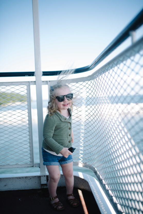 A young child wearing sunglasses, a green jacket, and denim shorts stands on a ferry deck. The childs hair is blown by the wind, and the background shows water and blue sky. by Justin Salem Meyer