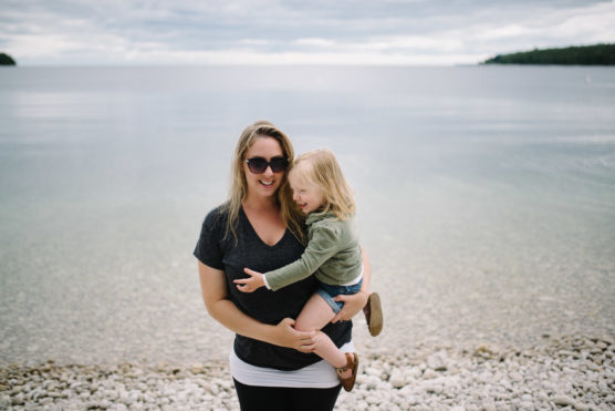 A woman wearing sunglasses holds a smiling young child by a calm, rocky lakeshore with clear water and a cloudy sky in the background. by Justin Salem Meyer