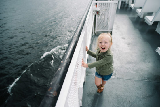 A young child with blonde hair, wearing a green jacket and shorts, smiles excitedly while standing on the deck of a boat near the water railing. The ocean is visible on the left side of the image. by Justin Salem Meyer