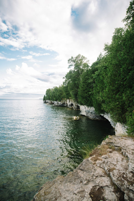 Rocky shoreline with cliffs covered in dense green trees next to calm, clear water under a partly cloudy sky. The landscape appears tranquil and natural, with the forest meeting the edge of the lake. by Justin Salem Meyer