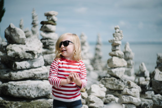A young child wearing sunglasses and a red-and-white striped shirt stands among stacked stone formations outdoors, smiling with the sea and sky in the background. by Justin Salem Meyer