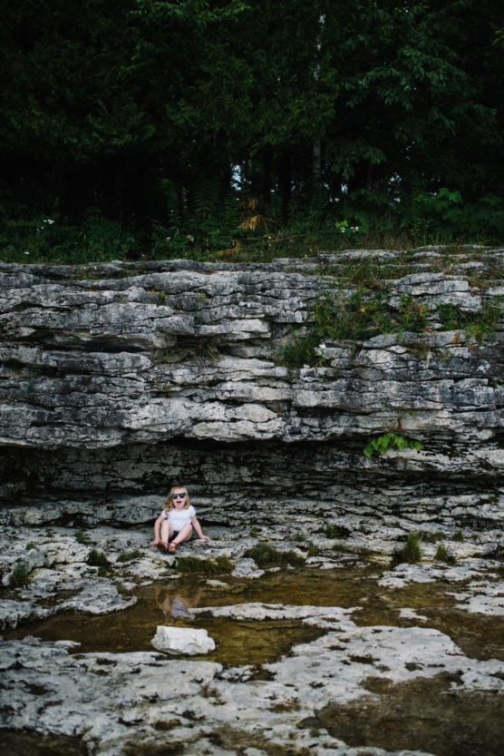 A young child wearing sunglasses sits on rocky ground near a small pool of water, with layered stone cliffs and dense green trees in the background. by Justin Salem Meyer