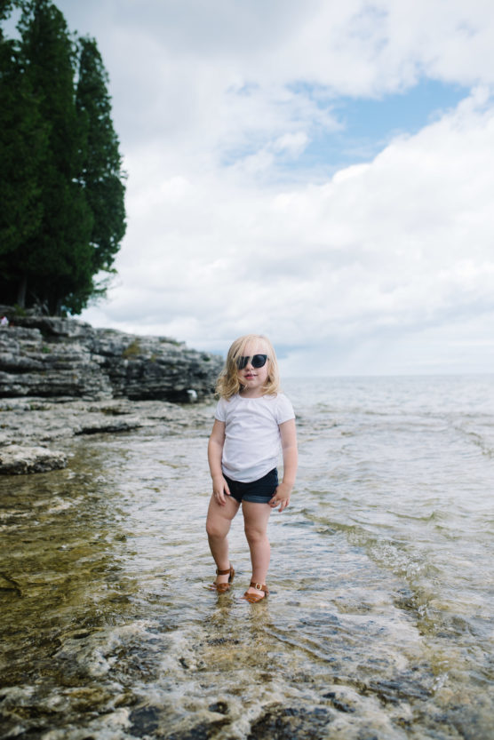 A young child wearing sunglasses, a white shirt, and shorts stands confidently on shallow rocky water near a shoreline with trees and a cloudy sky in the background. by Justin Salem Meyer