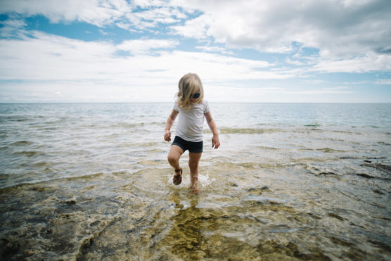 A young child wearing a white shirt, dark shorts, and sunglasses walks through shallow water on a rocky shore, with the ocean and a partly cloudy sky in the background. by Justin Salem Meyer