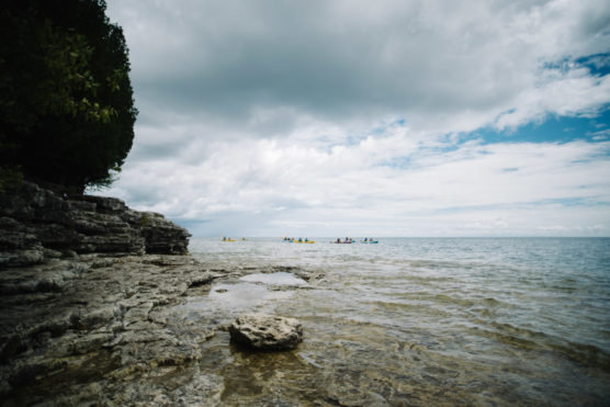 A rocky shoreline with clear water under a cloudy sky, trees on the left, and a group of kayakers paddling in the distance. by Justin Salem Meyer