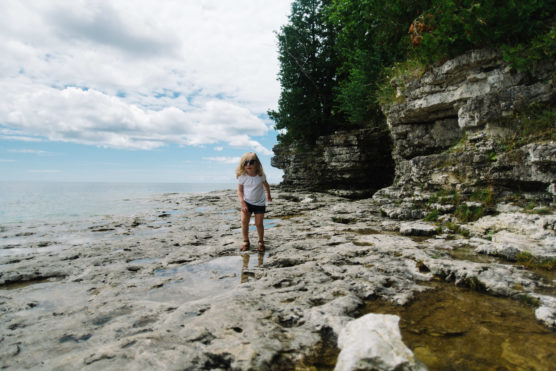 A young child with blonde hair stands on rocky terrain near a body of water, with layered cliffs and green trees in the background under a partly cloudy sky. by Justin Salem Meyer