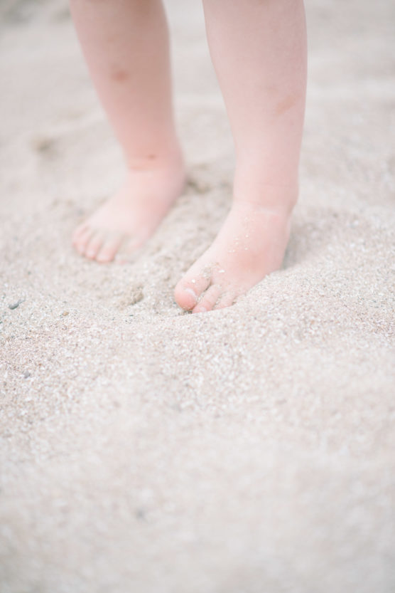 Close-up of a childs bare feet standing in light-colored sand, with only the legs and feet visible, suggesting a beach or sandbox setting. by Justin Salem Meyer