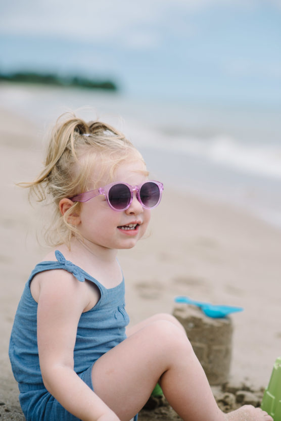 A young child in a blue outfit and purple sunglasses sits on a sandy beach, smiling with the ocean in the background and a toy shovel and sandcastle nearby. by Justin Salem Meyer