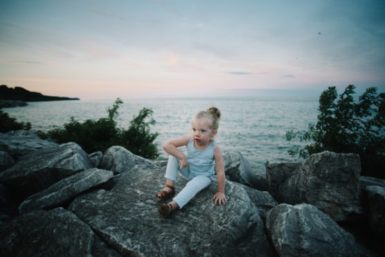 A young child with blond hair sits on large rocks near a calm body of water at sunset, with greenery and a soft pastel sky in the background. by Justin Salem Meyer