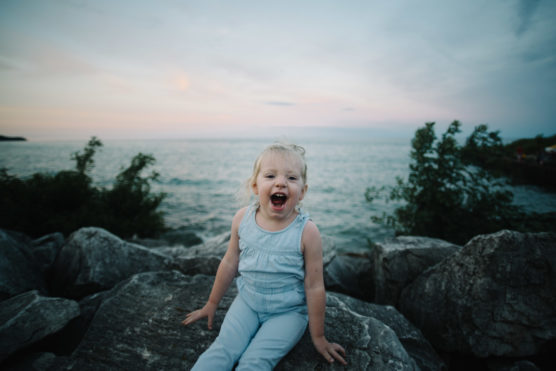 A young child in a light blue outfit sits on large rocks by the water, smiling joyfully with an open mouth. Trees frame the scene, and the sky appears cloudy at sunset. by Justin Salem Meyer