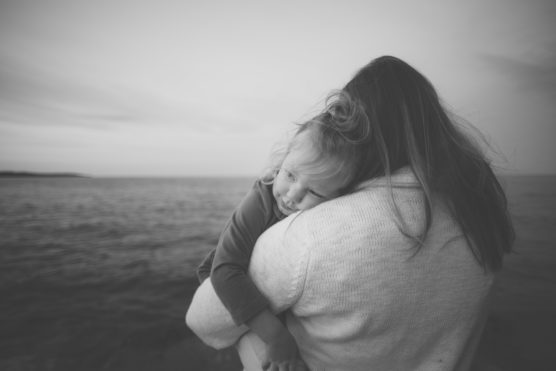 A black-and-white photo of a woman holding a young child by the sea. The child rests their head on the womans shoulder, looking content. Both have their backs to the camera, facing the water under a cloudy sky. by Justin Salem Meyer