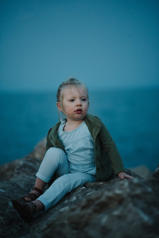 A young child with blonde hair, wearing a green jacket and light pants, sits on a large rock by the sea during dusk, with a calm blue ocean in the background. by Justin Salem Meyer