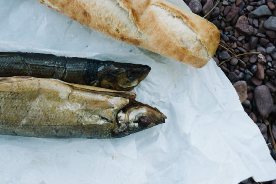 Two whole smoked fish lie on white parchment paper next to a partially visible baguette, with pebbles in the background. by Justin Salem Meyer