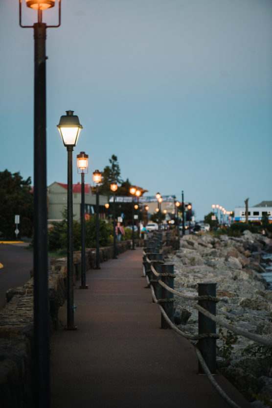 A paved walkway lined with lit street lamps runs alongside a rocky shoreline at dusk; buildings and trees are visible in the background. by Justin Salem Meyer