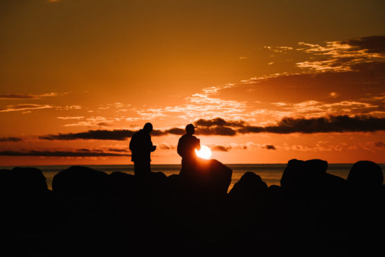 Two people stand on rocks by the water, silhouetted against an orange sunset sky with scattered clouds. The sun is low on the horizon, casting a warm glow over the scene. by Justin Salem Meyer