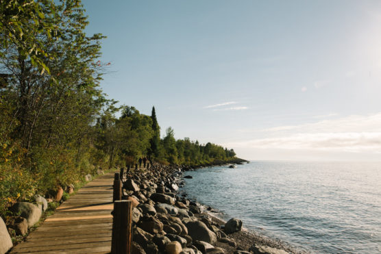 A wooden boardwalk runs along a rocky shoreline bordered by green trees, with calm blue water on the right and a bright, clear sky overhead. by Justin Salem Meyer
