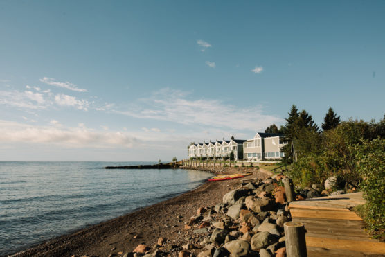 A rocky shoreline with calm water, a sandy beach, and a row of modern white buildings facing the water, surrounded by trees under a clear blue sky. by Justin Salem Meyer