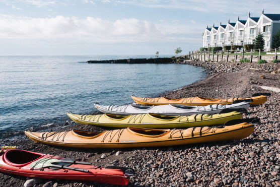 Several colorful kayaks rest on a rocky shoreline near calm water, with a row of white, multi-story buildings and a partly cloudy sky in the background. by Justin Salem Meyer
