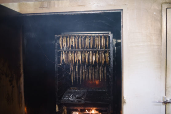 Rows of fish hang inside a large smokehouse with smoke rising and a small fire burning below, used for traditional fish smoking. by Justin Salem Meyer