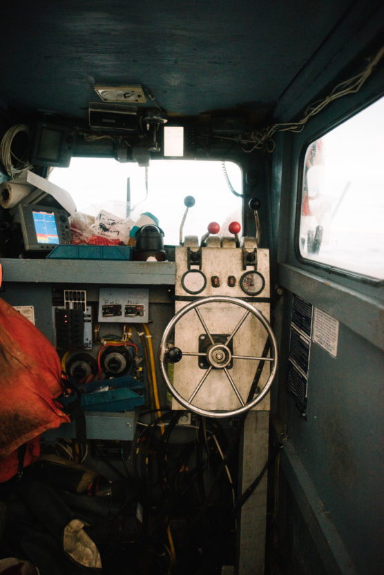 The interior of a small boats control cabin, featuring a metal steering wheel, various gauges and levers, and equipment scattered around. Light enters through the side window. by Justin Salem Meyer