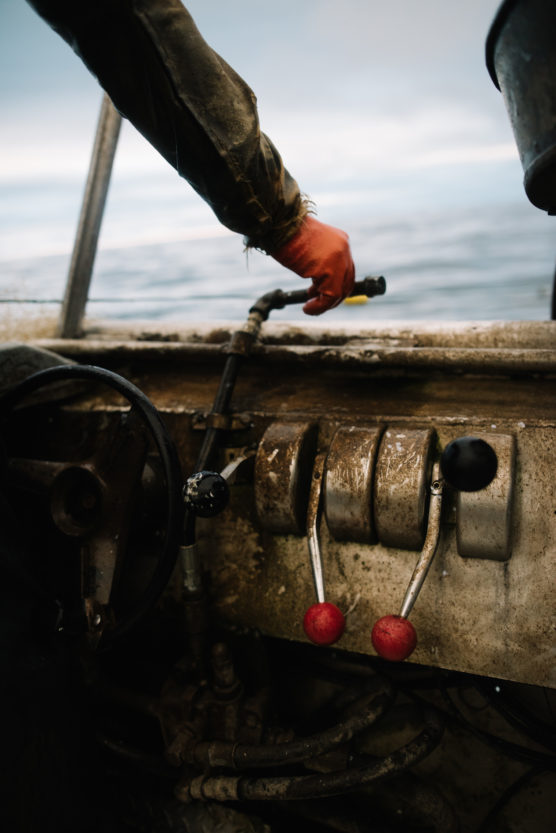 A person wearing an orange glove operates the controls of a weathered boat, with two red-handled levers visible near a steering wheel, and the sea in the background. by Justin Salem Meyer