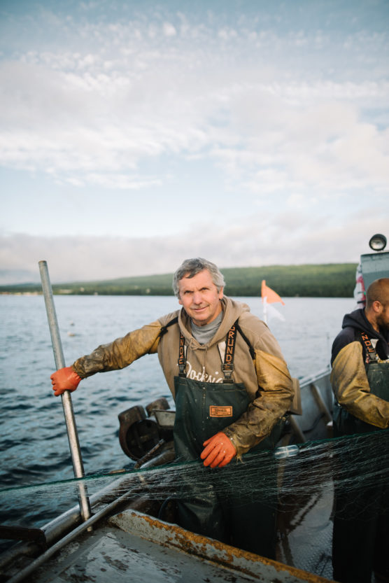 A fisherman in waterproof overalls and orange gloves stands on a boat holding a fishing net, with water and a distant shoreline visible under a cloudy sky. by Justin Salem Meyer