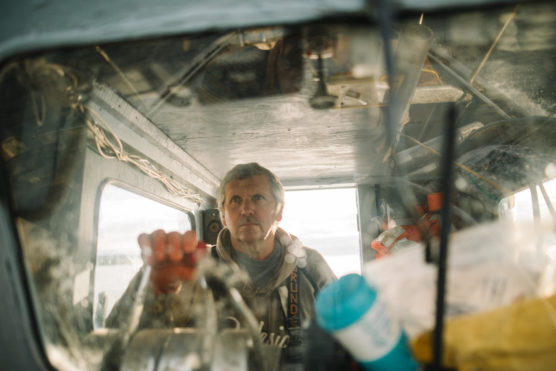 A man steers a boat, viewed through a dirty window cluttered with various objects, looking focused as he navigates. The interior appears worn, with ropes and equipment visible around him. by Justin Salem Meyer