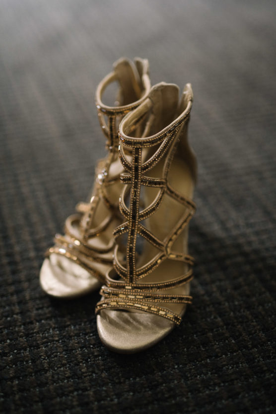 A pair of elegant, gold high-heeled sandals adorned with sparkling rhinestones, displayed on a dark, textured carpet. by Justin Salem Meyer