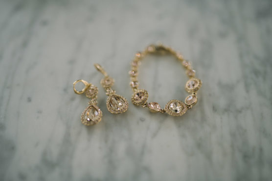 A pair of gold drop earrings and a matching gold bracelet with clear gemstones are displayed on a gray marble surface. by Justin Salem Meyer