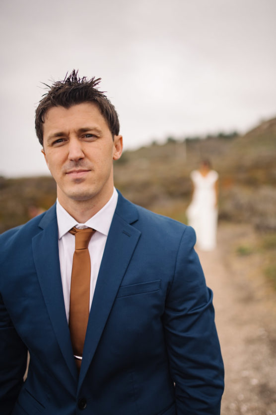 A man in a blue suit and brown tie stands outdoors, looking at the camera with a neutral expression. In the blurred background, a woman in a white dress stands on a path amidst natural scenery. by Justin Salem Meyer