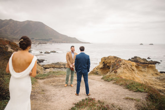 A bride in a white dress approaches two men—one in a tan jacket, the other in a blue suit—standing on a coastal cliff overlooking the ocean, with mountains and cloudy skies in the background. by Justin Salem Meyer