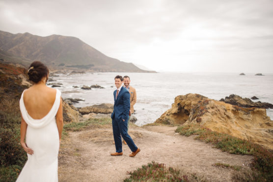 A bride in a white dress approaches two men, one in a blue suit and one in a tan suit, standing on a rocky coastal path with waves and mountains in the background under a cloudy sky. by Justin Salem Meyer