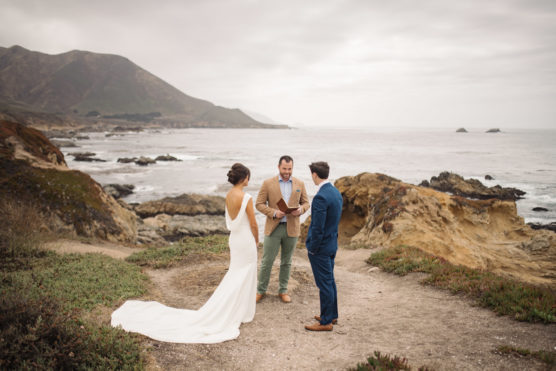 A bride in a white dress and a groom in a blue suit stand on a rocky coastal cliff, facing an officiant as they exchange vows with the ocean and mountains in the background under a cloudy sky. by Justin Salem Meyer