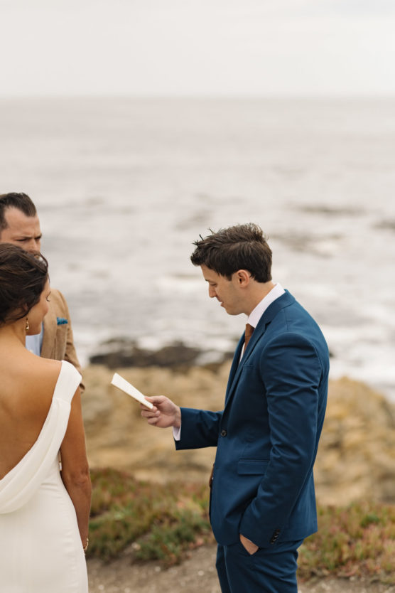 A man in a blue suit reads vows from a card to a woman in a white dress with an open back, standing outdoors on a rocky coastline with the ocean in the background. by Justin Salem Meyer