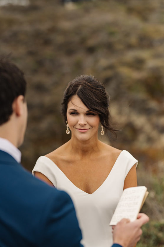 A bride in a white dress with drop earrings smiles softly at a man in a blue suit who is holding a handwritten note, standing outdoors with a blurred natural background. by Justin Salem Meyer