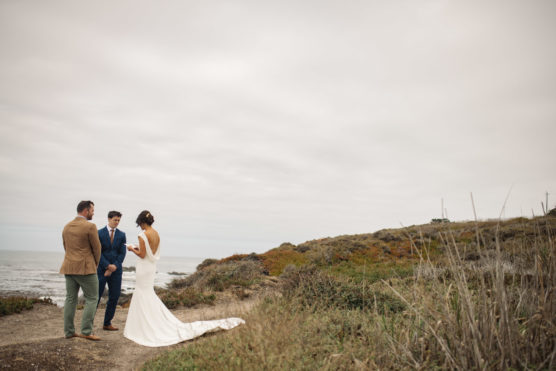 A bride in a white dress stands with two men on a grassy, coastal cliff overlooking the ocean under a cloudy sky, appearing to exchange vows in an intimate outdoor ceremony. by Justin Salem Meyer