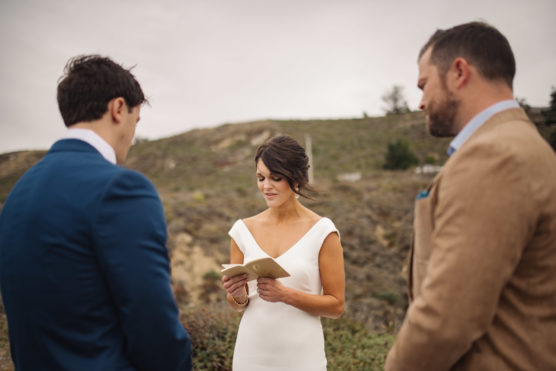 A bride in a white dress reads from a booklet while standing outdoors between two men in suits, with a hilly landscape in the background. by Justin Salem Meyer