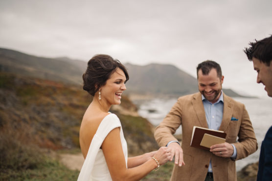 A bride in a white dress smiles as she exchanges rings with her partner during an outdoor wedding ceremony, with an officiant holding a book and mountains and ocean in the background. by Justin Salem Meyer